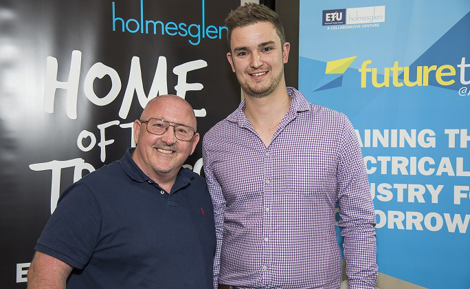 Electrical apprentice stands next to employer and smiles to camera.